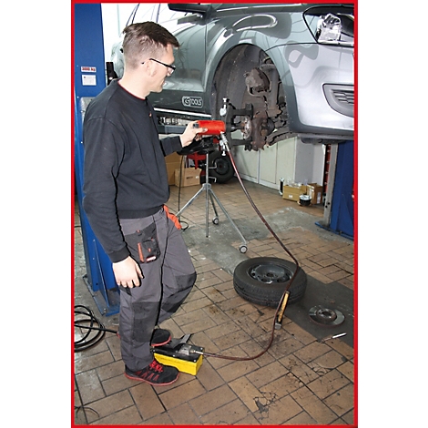 Ein Mechaniker repariert ein Auto in einer Werkstatt. Er benutzt ein Werkzeug, das an eine Pumpe angeschlossen ist. Ein Reifen liegt daneben.